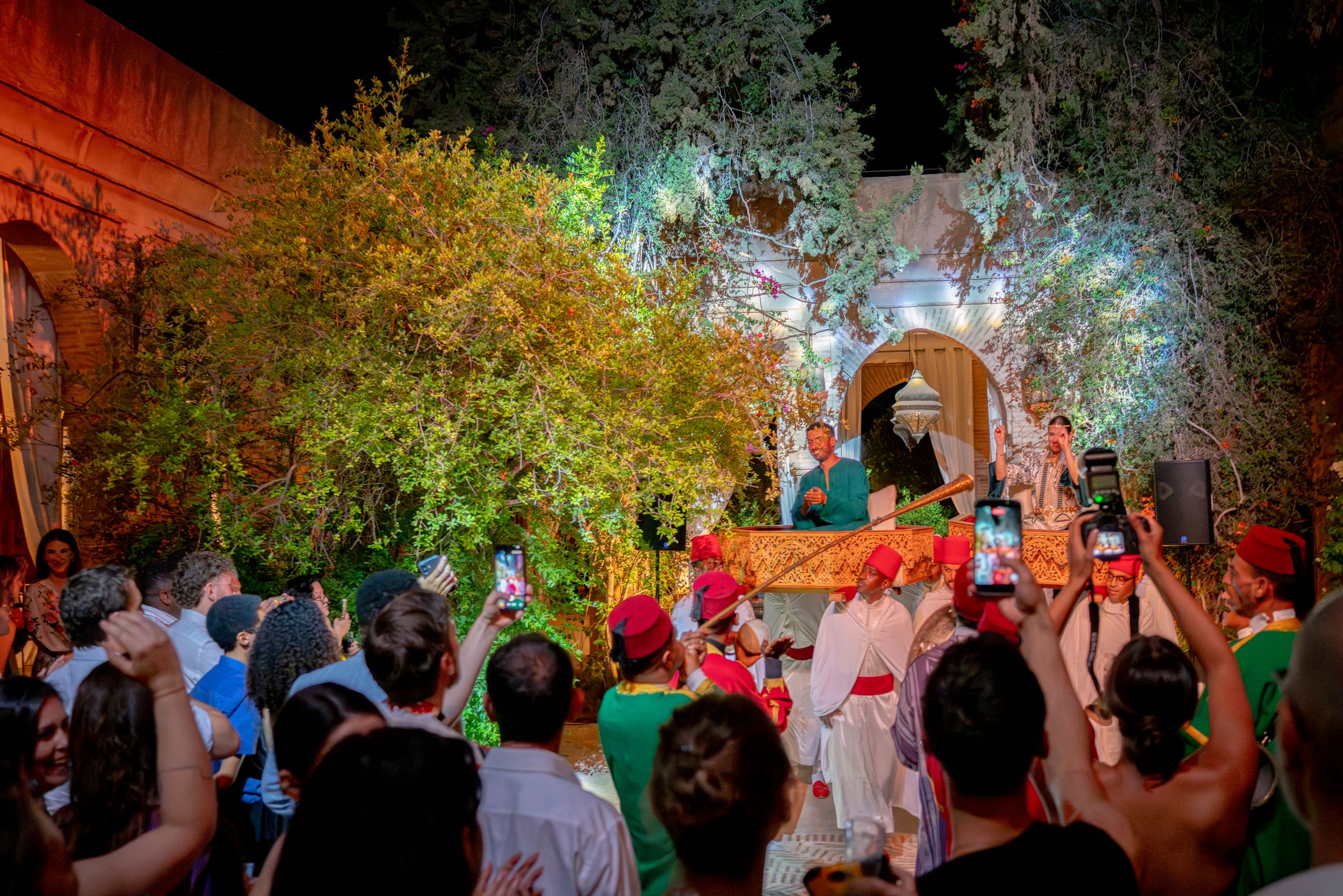 Festive nighttime wedding procession with crowds gathering around illuminated floral display outdoors.