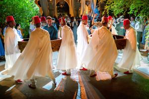 Men in white robes and red fez hats perform a traditional Moroccan dance ceremony indoors.