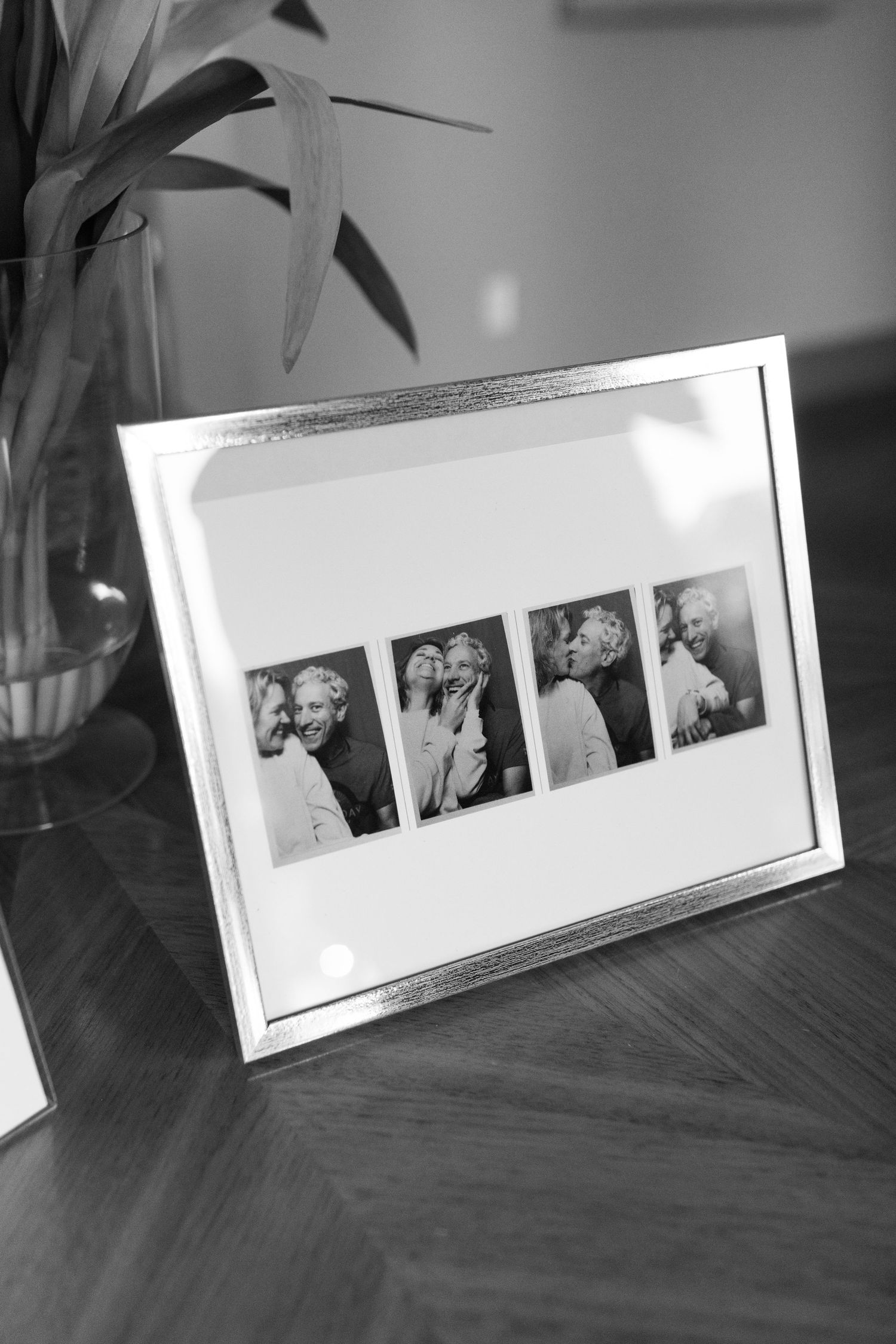 Black and white photo frame displaying four small portrait photos on a wooden table beside a glass vase with leaves.