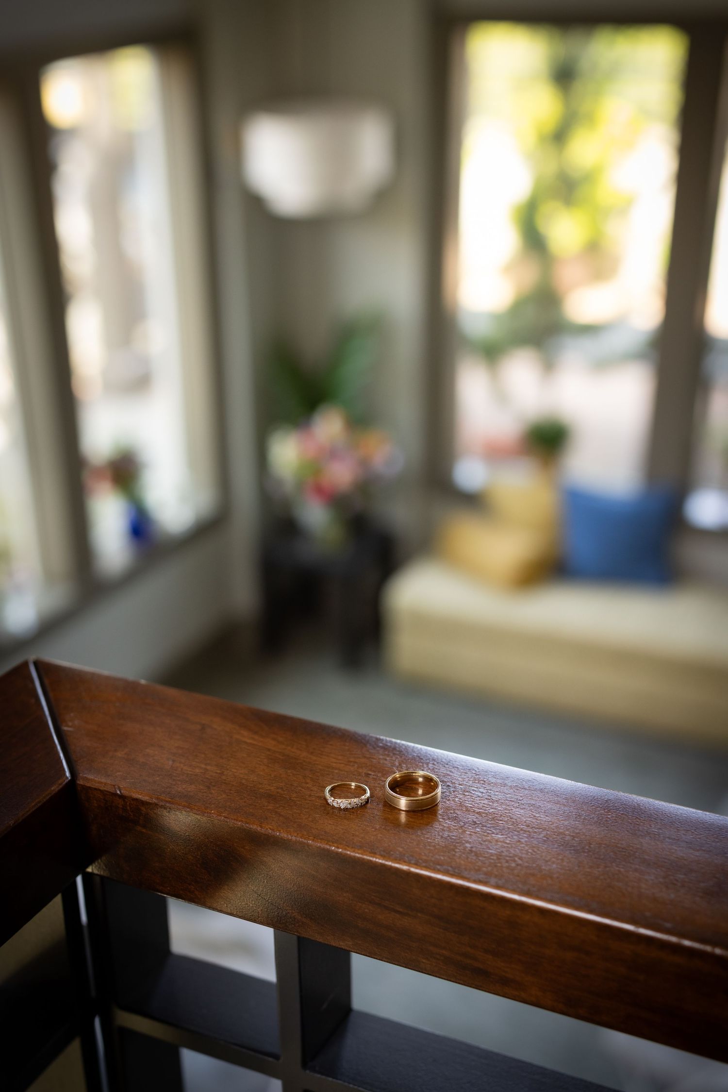 Two gold wedding rings resting on a wooden chair rail in a softly lit room with windows in the background.