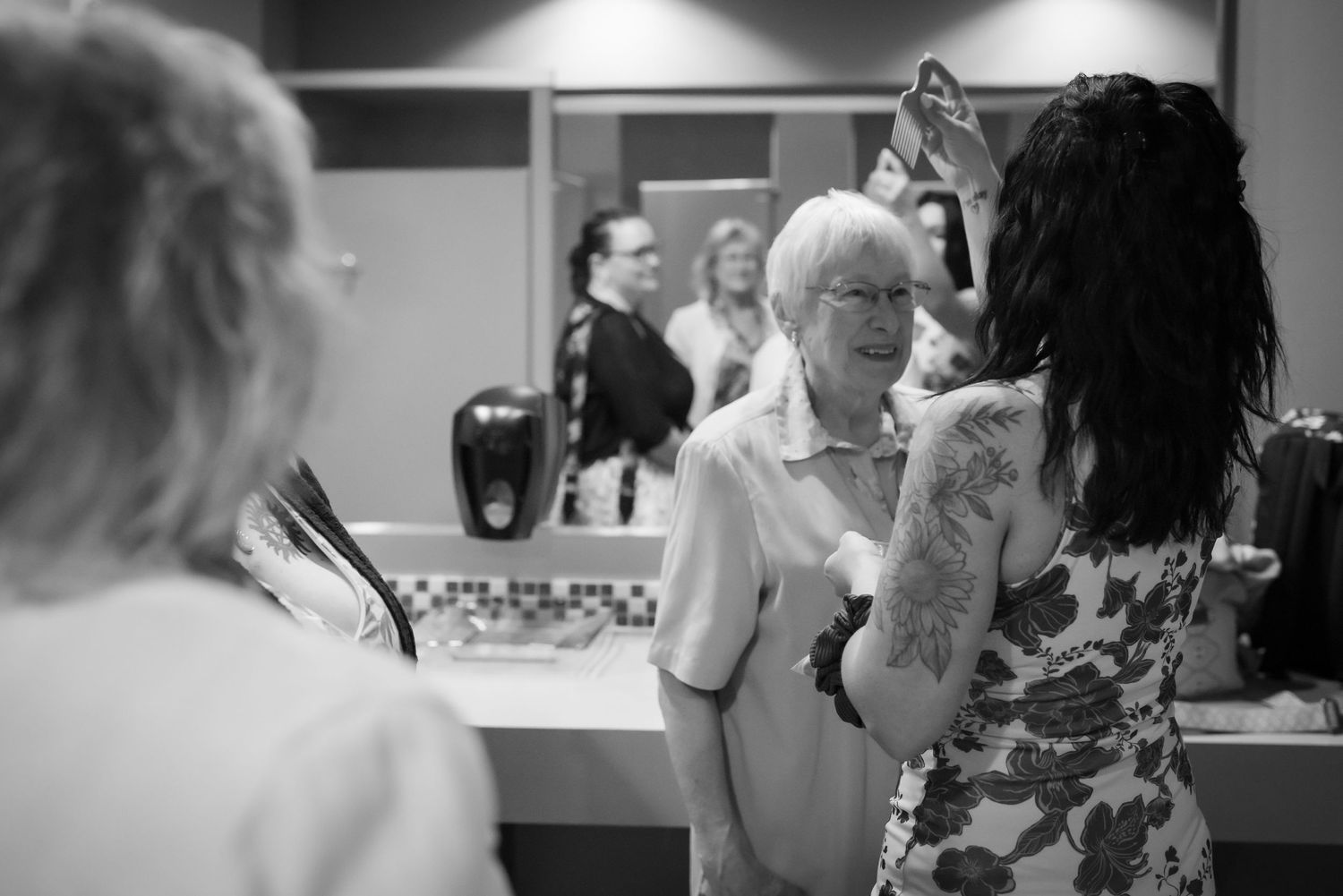 Black and white photo of women in a salon, one with tattoos styling an older woman's hair near a mirror.