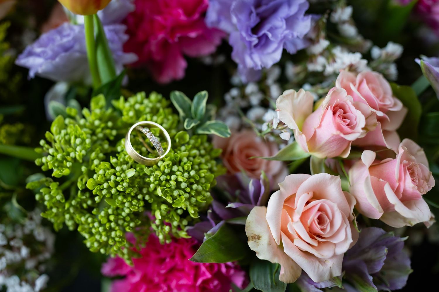 Wedding rings resting on a lush bouquet of pink roses, purple hyacinths, and vibrant green flowers.
