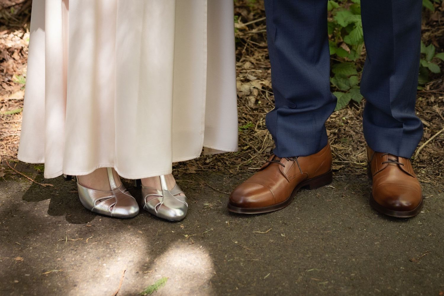 Close-up of wedding couple's feet, bride in silver shoes with white gown, groom in brown leather dress shoes and blue suit.