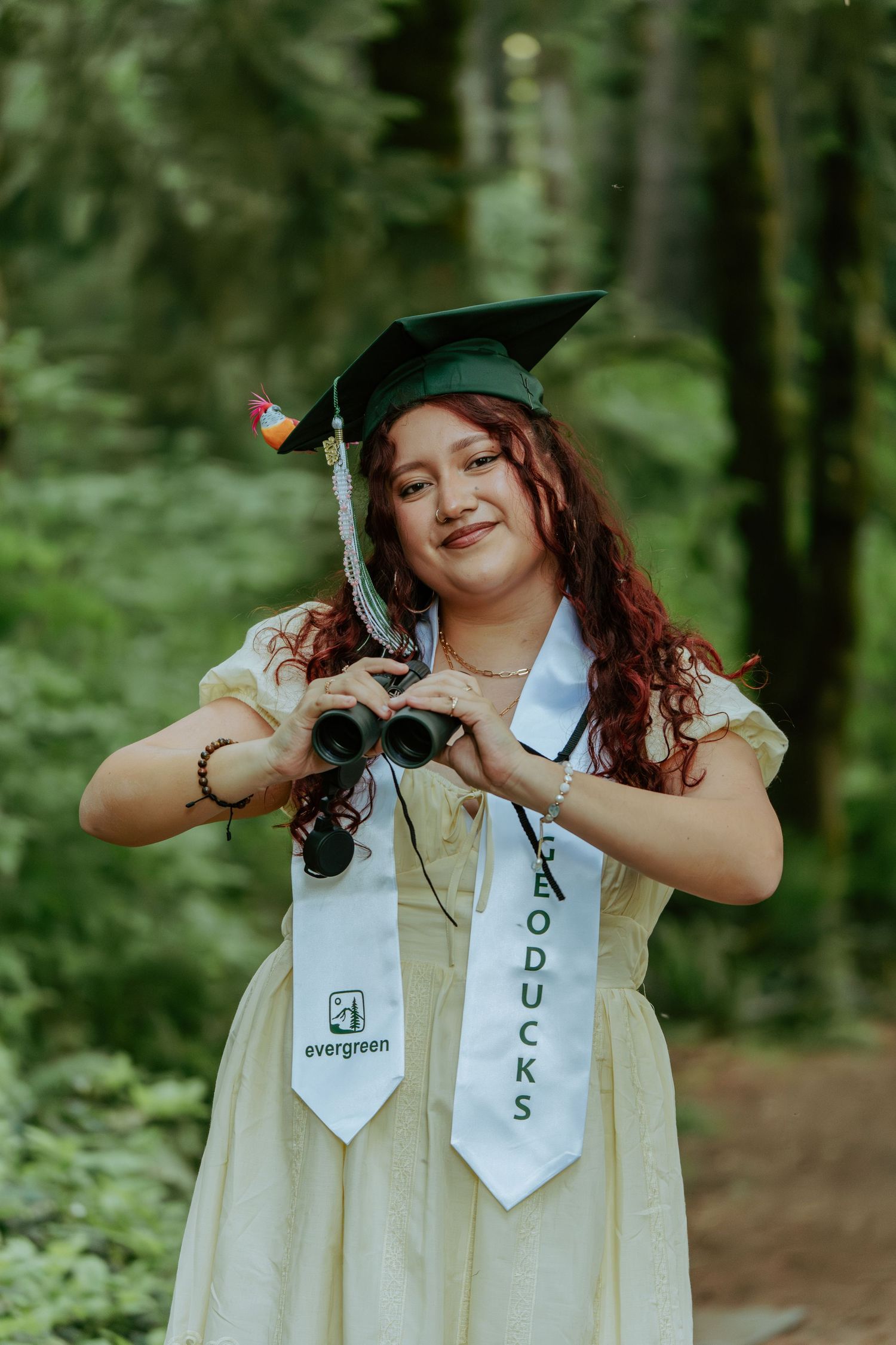 Smiling graduate holds binoculars forming a heart shape, wearing green cap and Geoducks stole.