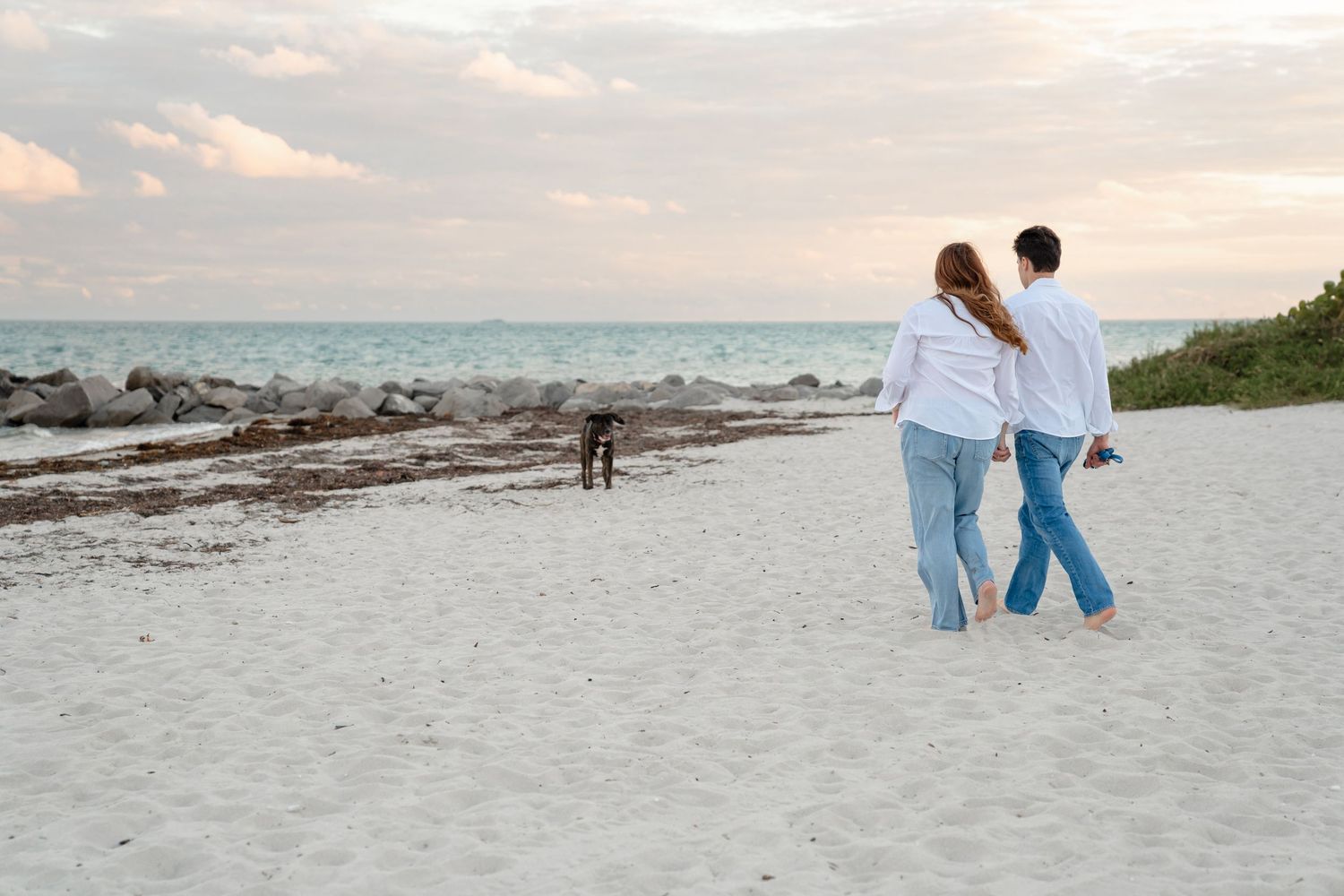 Couple walking barefoot on sandy beach at sunset with a dog near rocky shoreline and turquoise ocean.