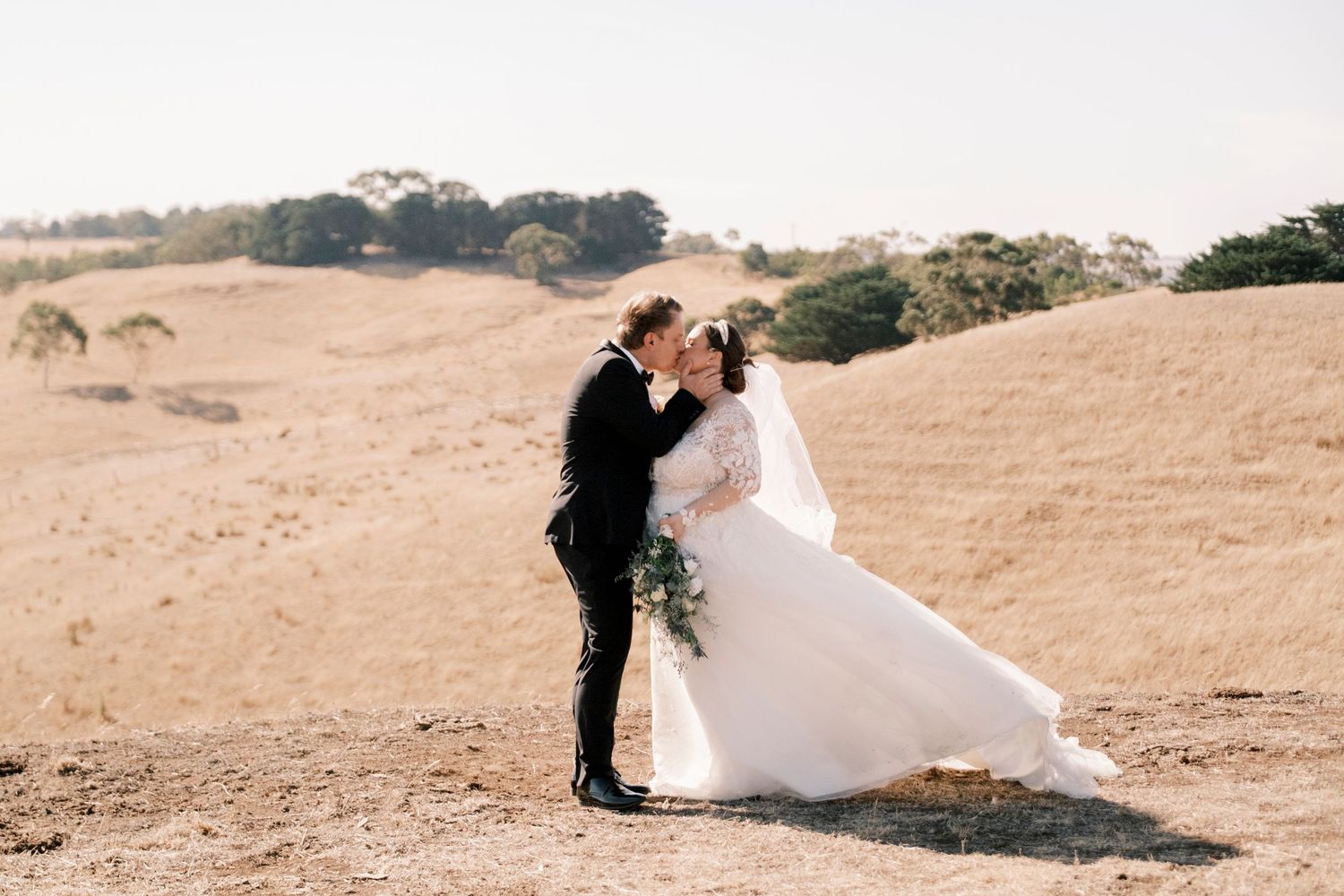 A wedding couple embracing on a golden hillside with flowing white dress in the wind.