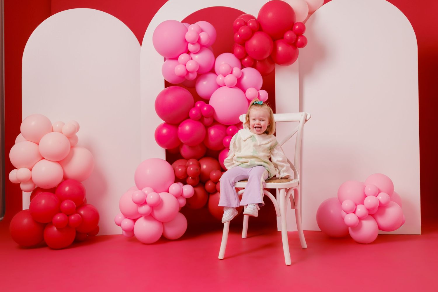 A happy toddler sits on a white chair surrounded by pink and red balloon decorations against a bright pink backdrop.