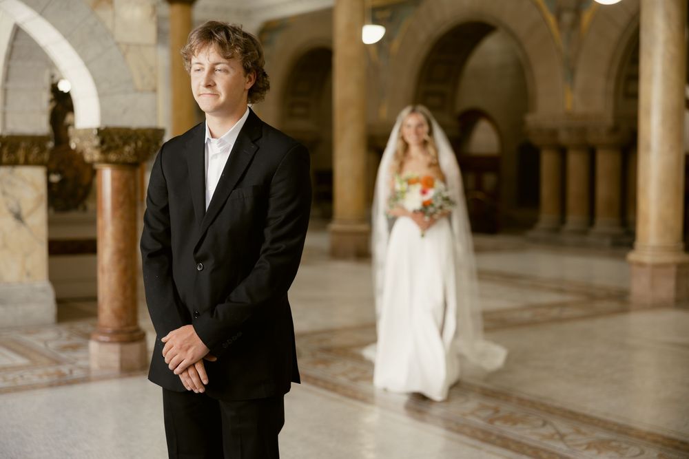 Groom in black suit stands in marble hall as bride in white gown and veil approaches from behind.