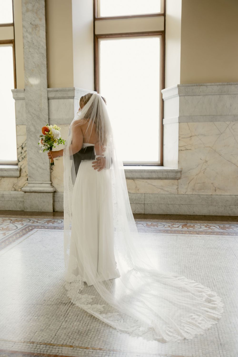 Bride in white wedding gown and long lace-trimmed veil holds colorful bouquet near sunlit window.