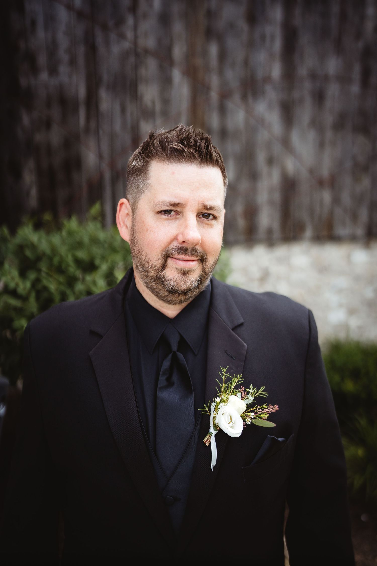 A groom in a black suit with a white boutonniere poses outdoors for a formal wedding portrait.