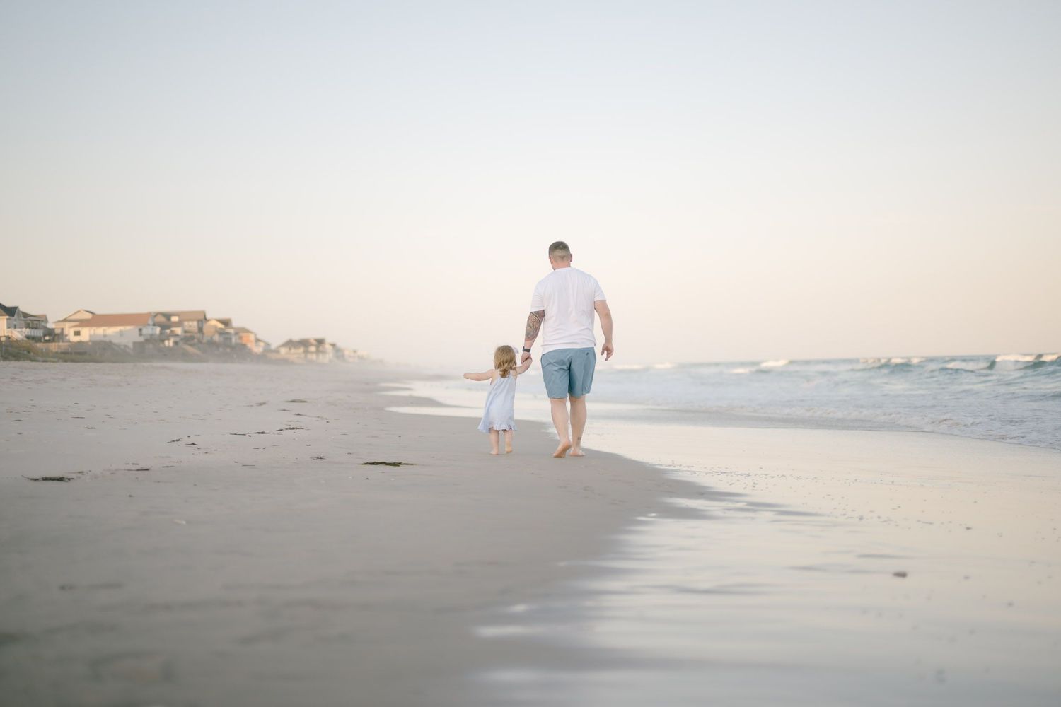 A father and young child walk hand-in-hand along a serene beach at sunset, with gentle waves and coastal homes in the background.