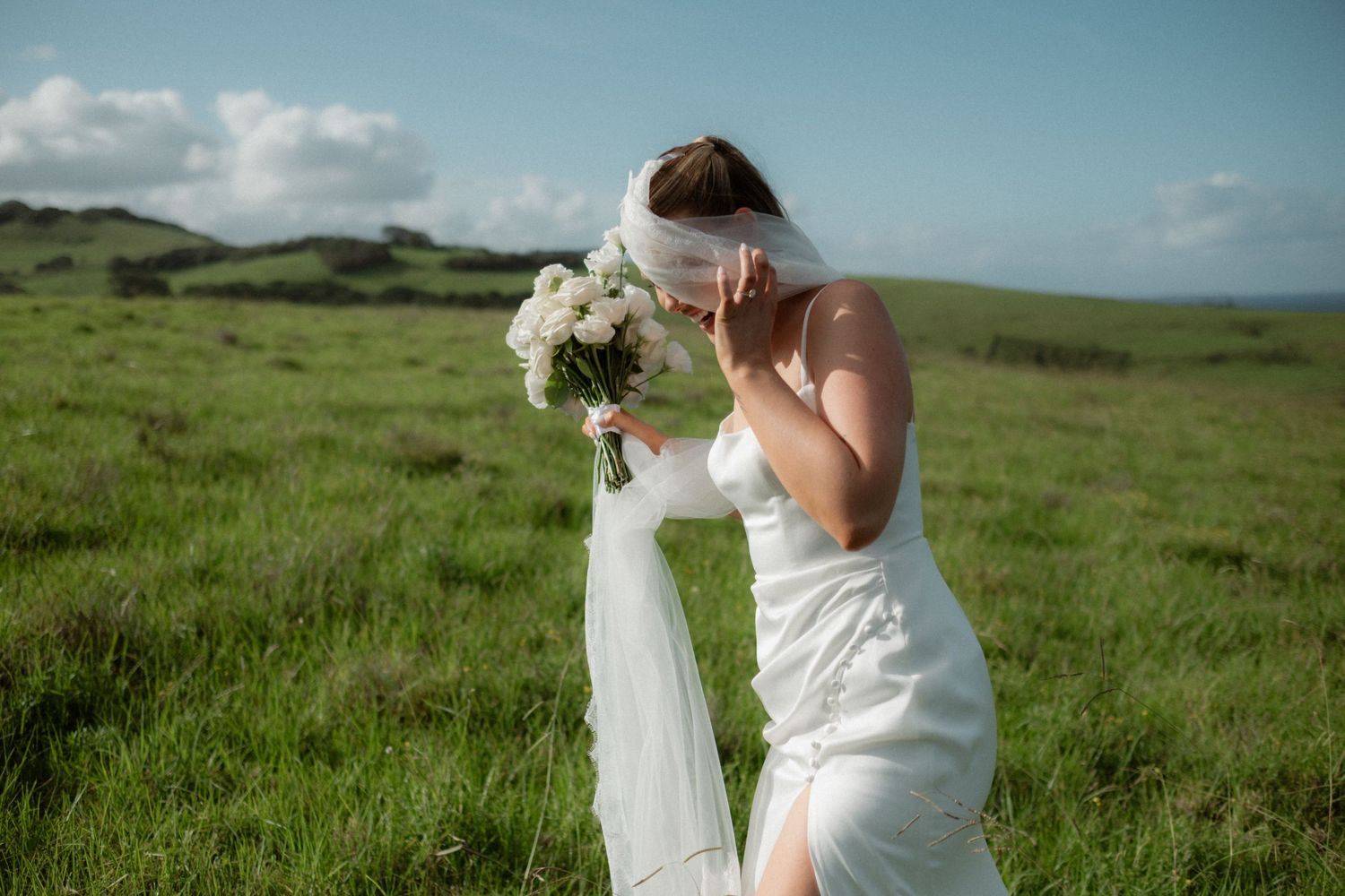 Emotional bride in white gown holding bouquet of white flowers in lush green field on her wedding day.