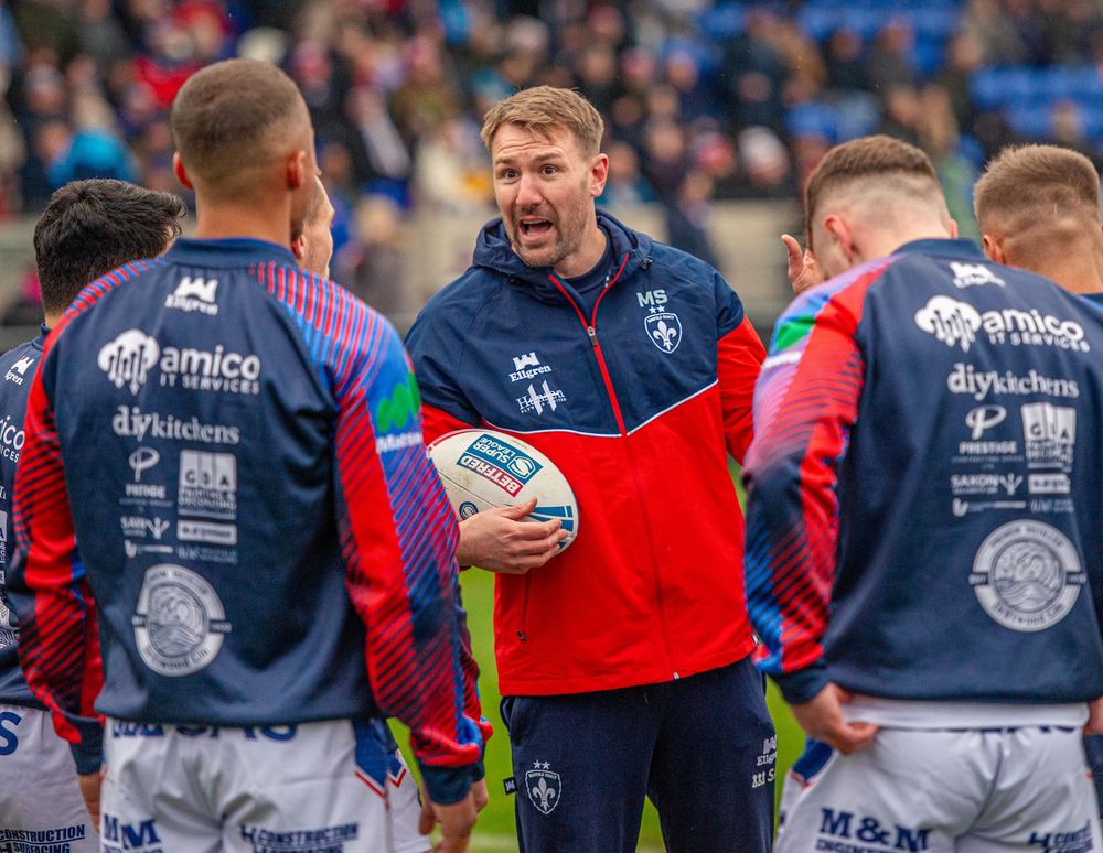 Rugby league coach addresses players in red and blue kits holding a match ball before a game.