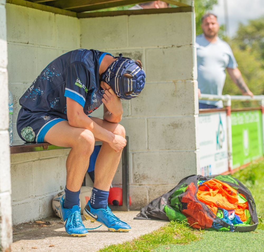 Dejected rugby player sits head in hands on substitutes bench during a sunny match day.