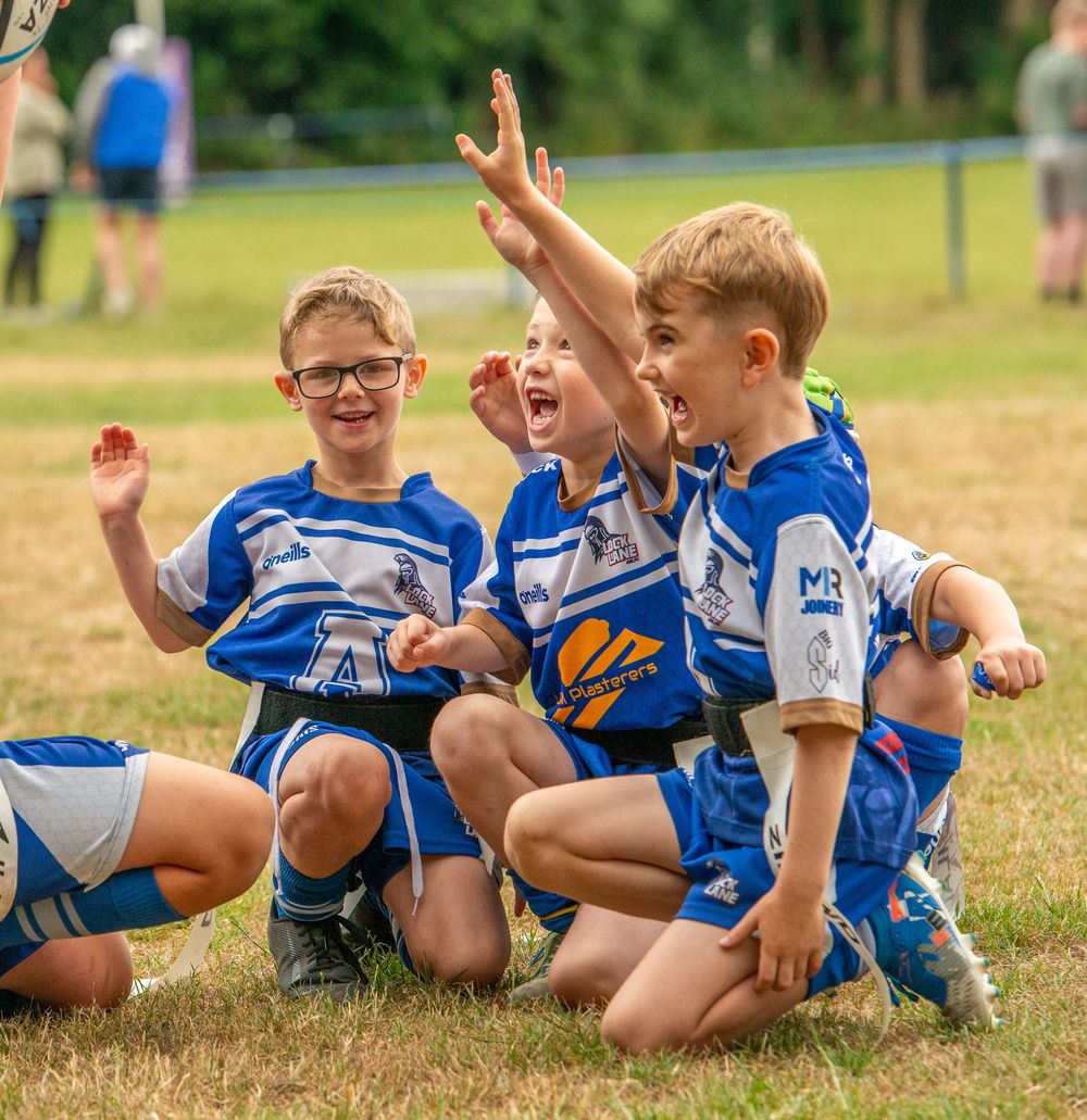Three joyful young boys in blue rugby league kits celebrate together on the grass during a match.