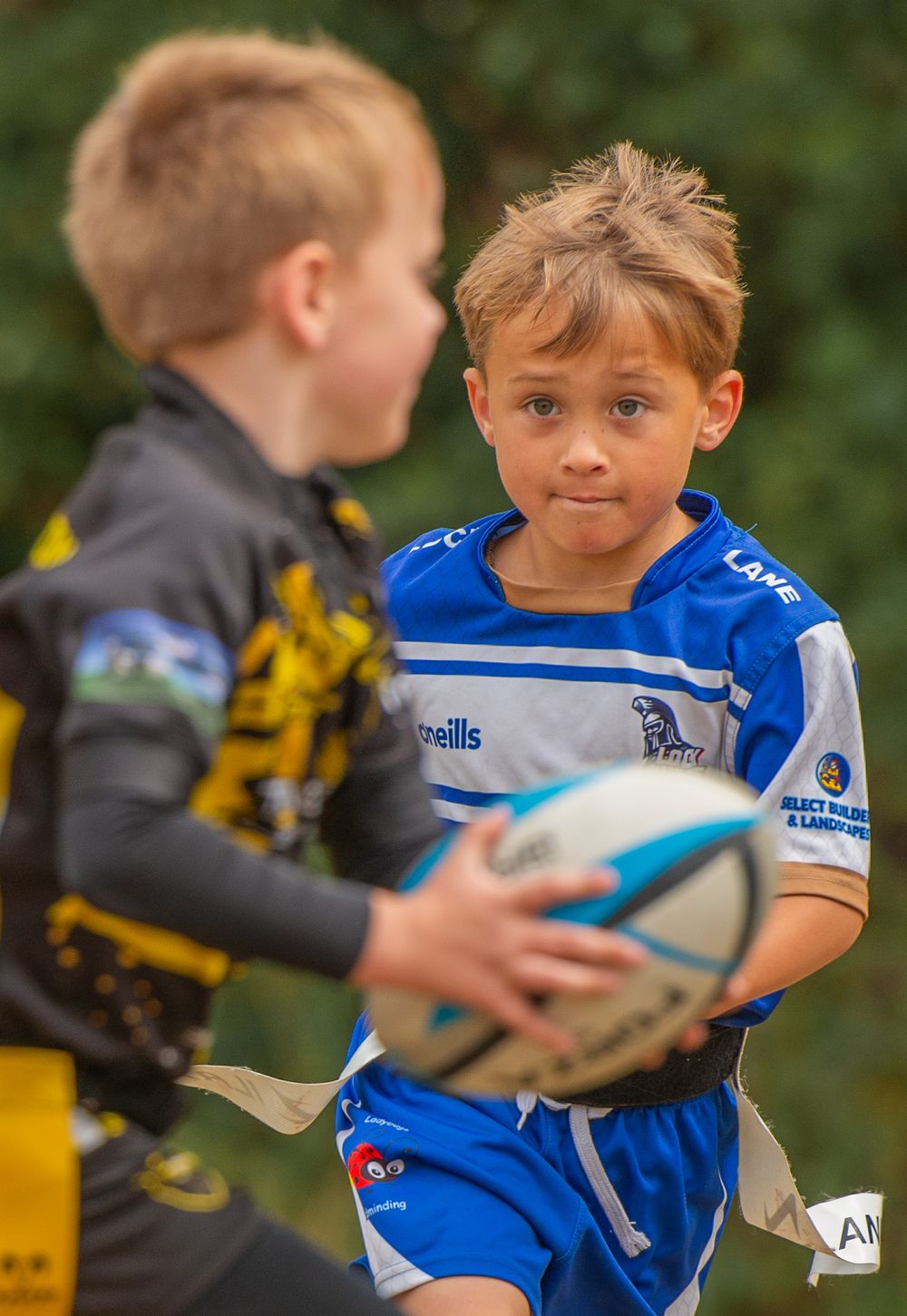 Two young boys compete in a junior rugby league match, one in blue carrying the ball.