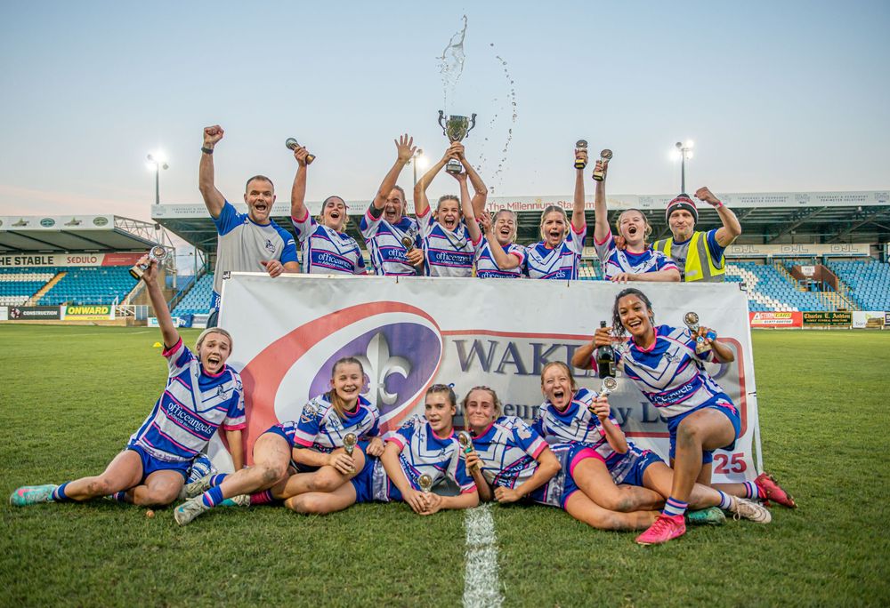 Womens rugby league team celebrates winning the Wakefield Trinity Cup trophy on the pitch.