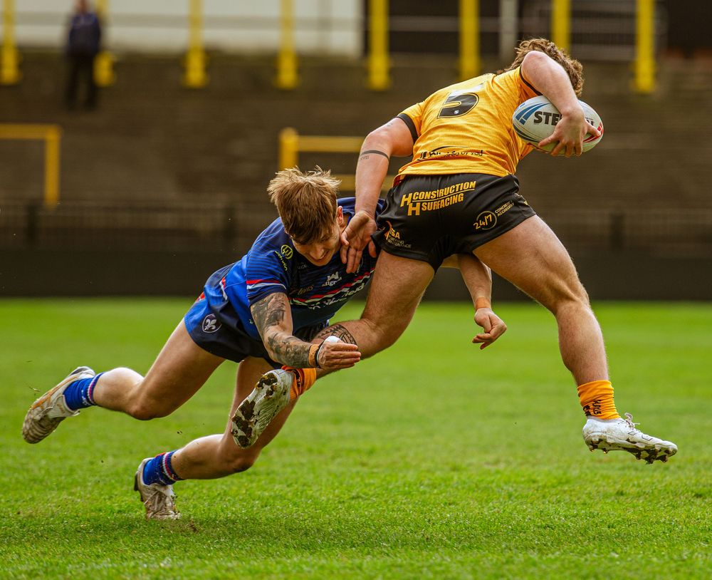 Rugby league tackle as player in blue brings down opponent in yellow kit during competitive match.