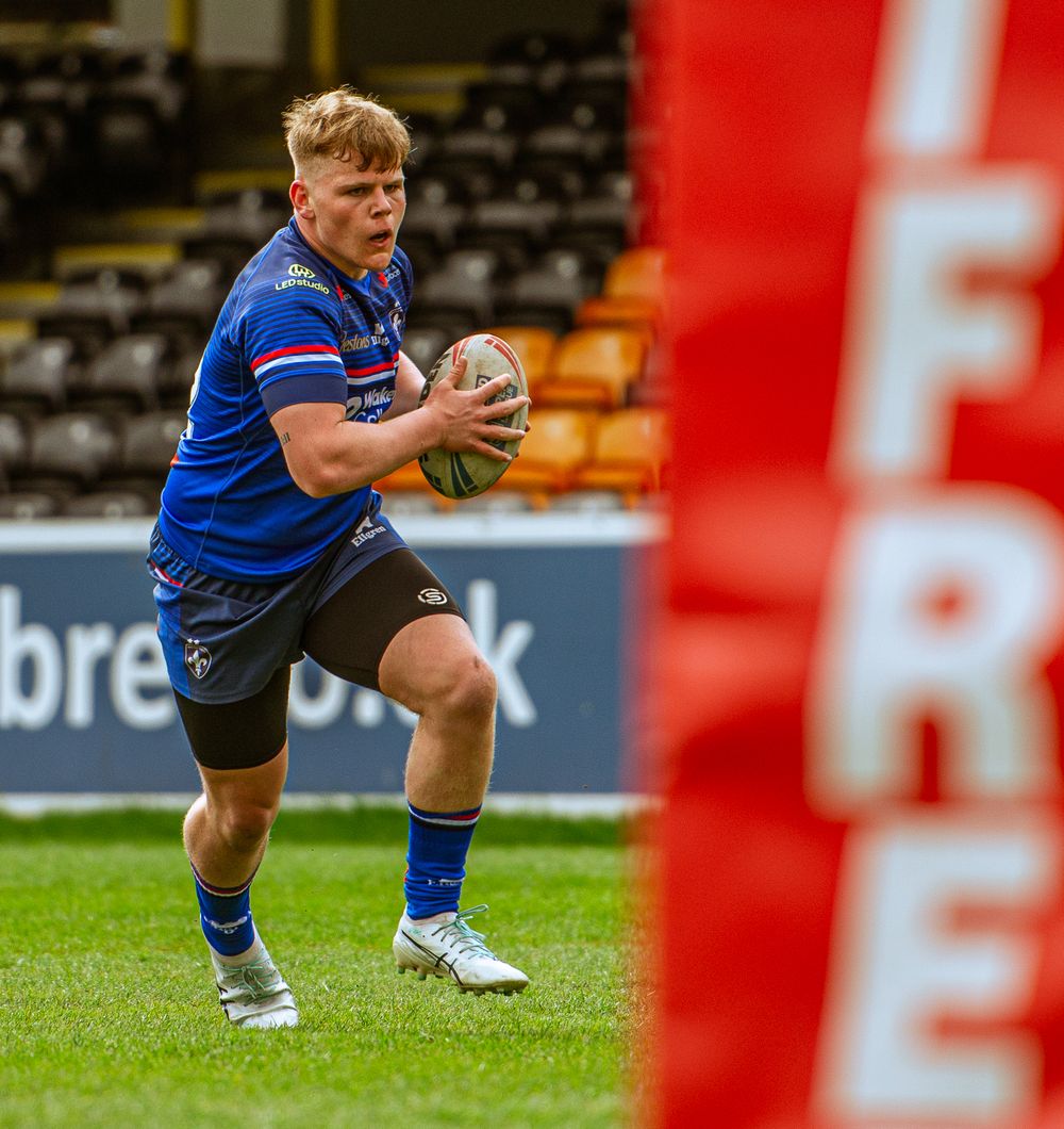 Young rugby league player in blue carries the ball during a junior match at a stadium.