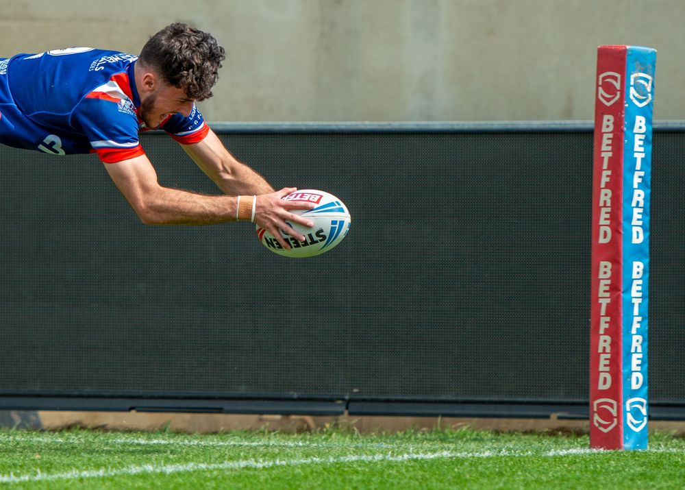 Rugby league player dives over the try line near the post in a blue and red kit during a Betfred match.