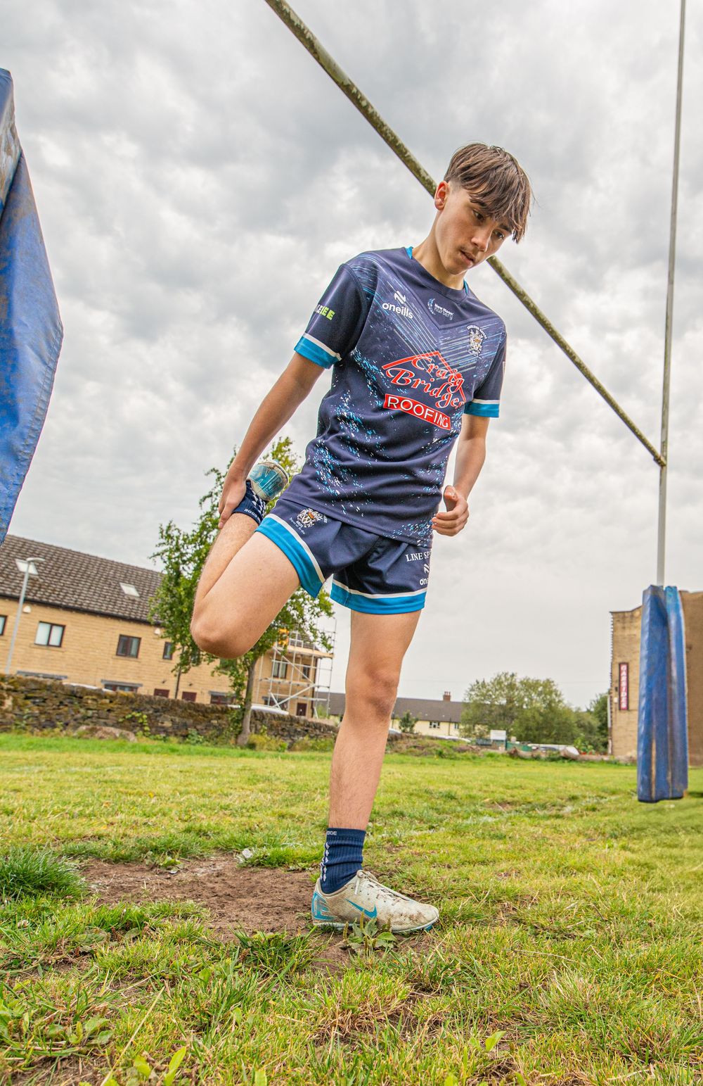 Young rugby league player in navy blue kit stretches near goalposts on a grass field under cloudy skies.