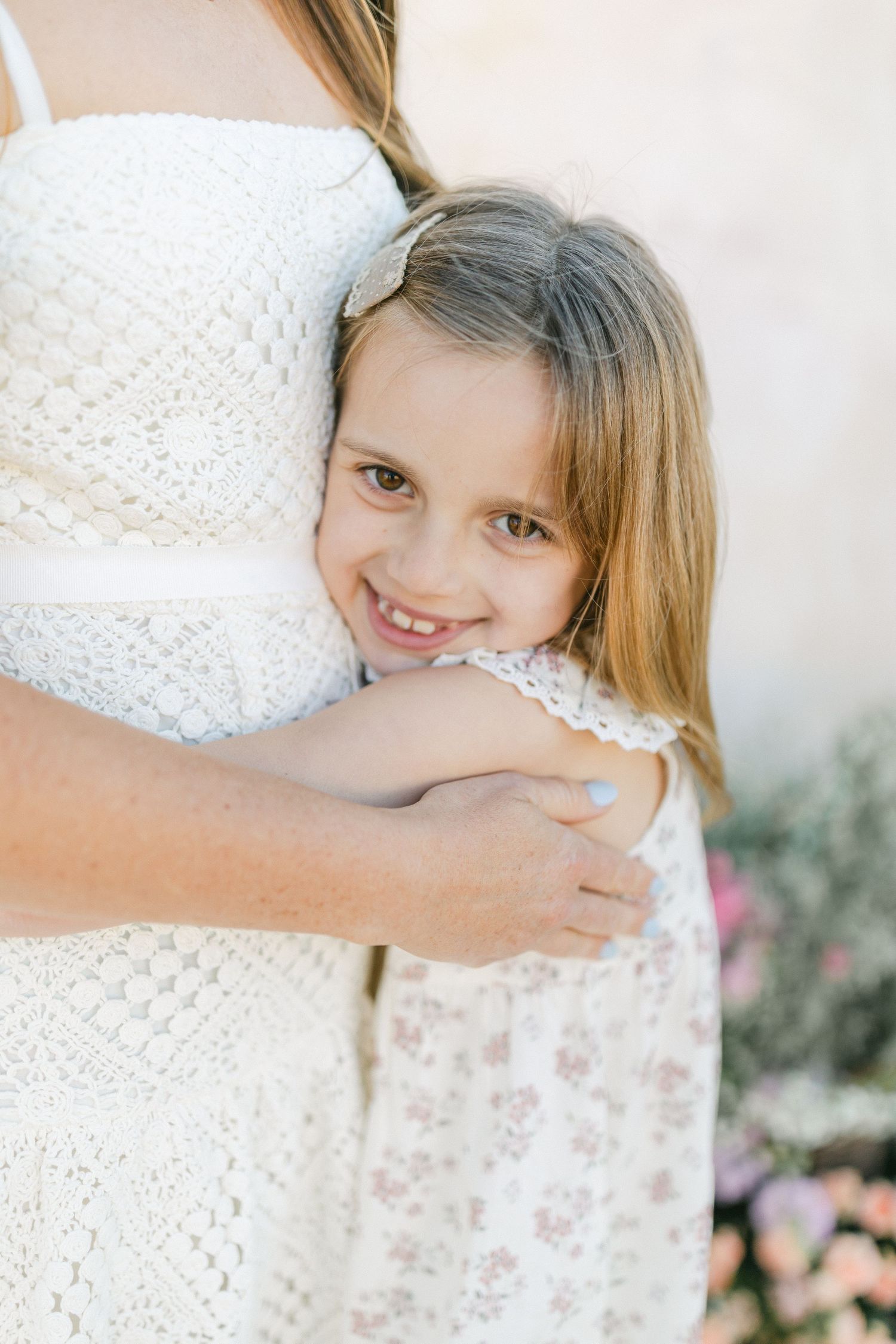 A young girl in a floral dress smiles while hugging a woman in a white lace dress outdoors.