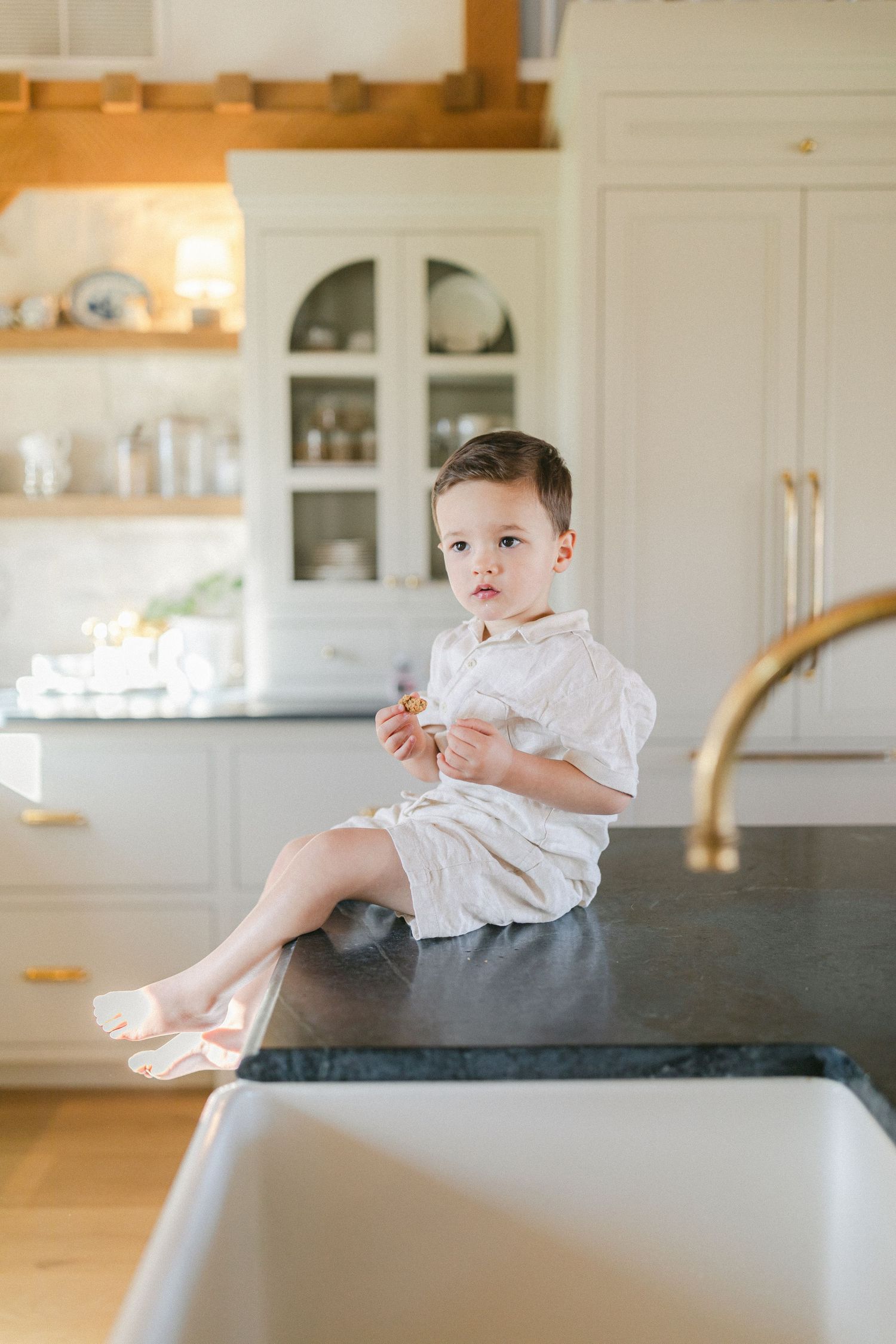 A happy toddler boy in a white linen outfit sits on a dark kitchen counter, smiling and holding a snack.