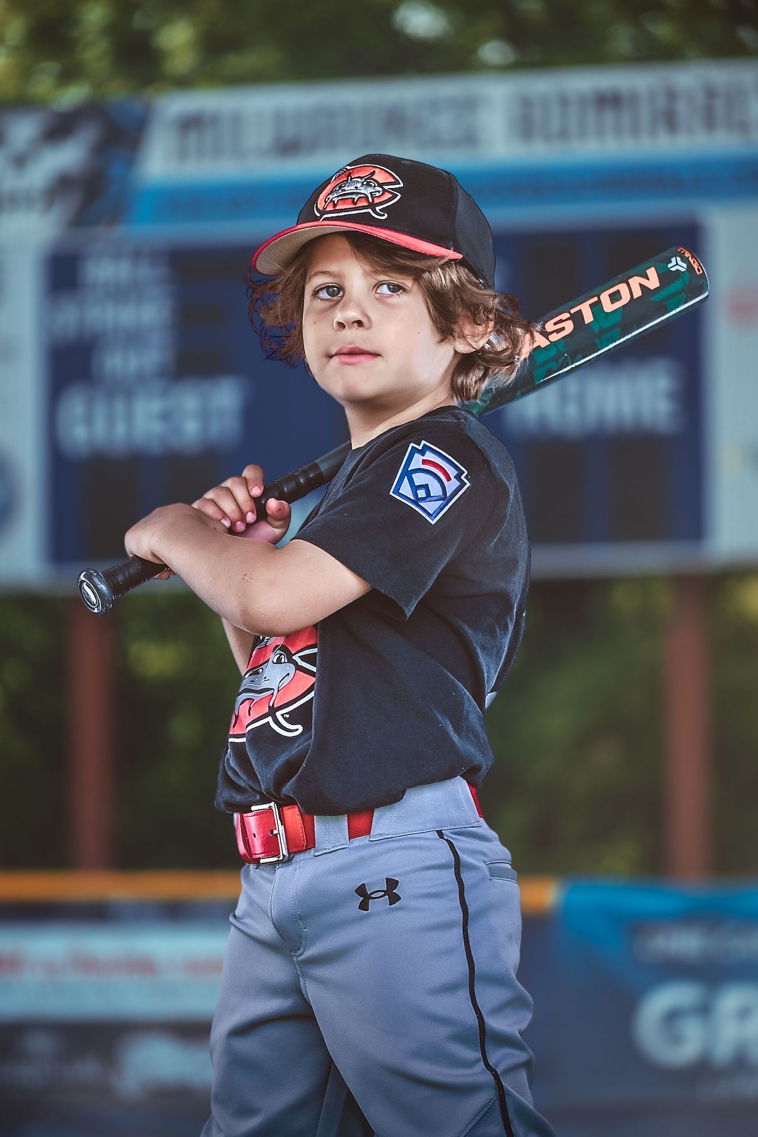 Young baseball player in uniform poses with Easton bat on shoulder at outdoor ballpark scoreboard backdrop.