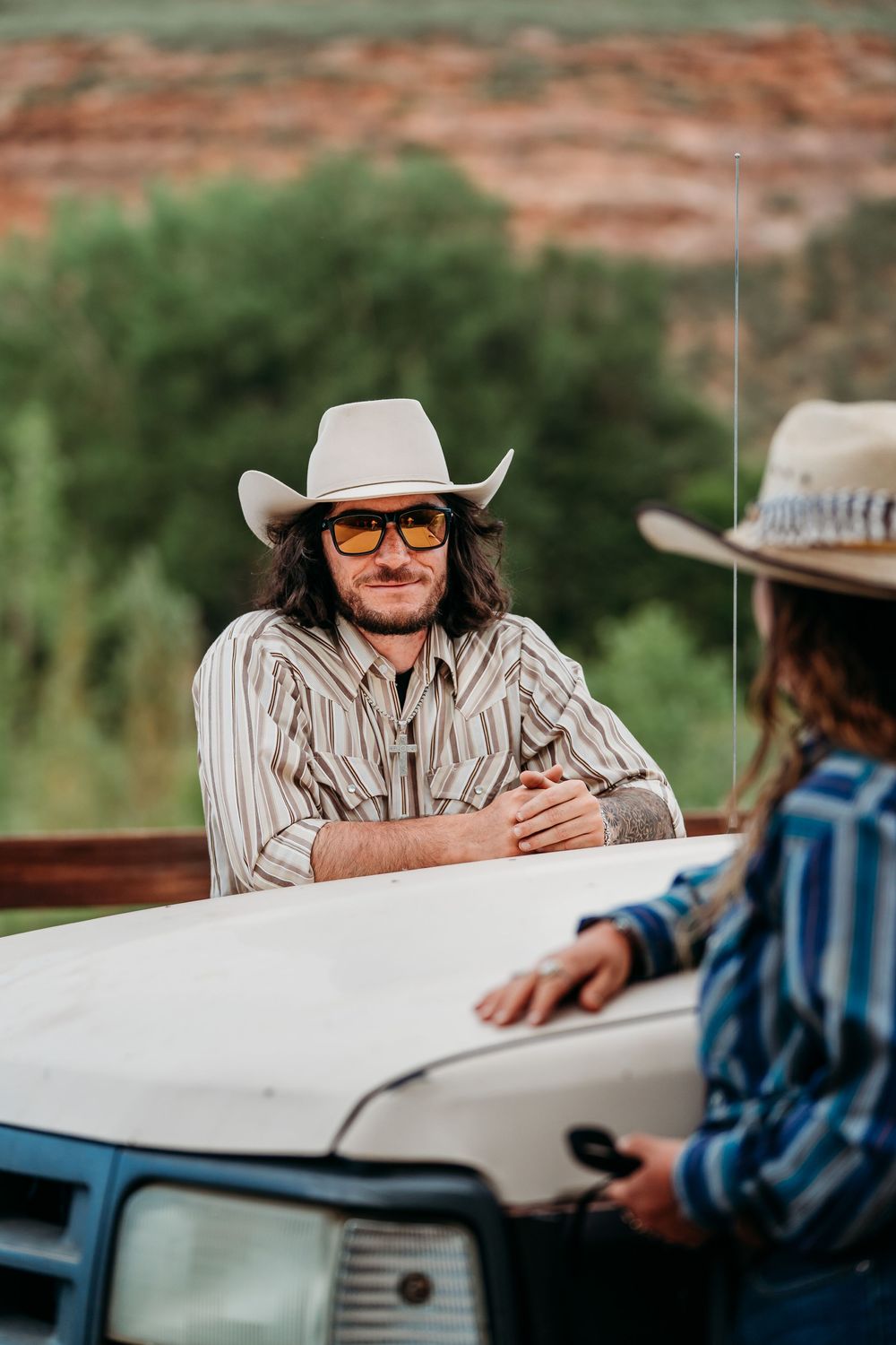 Two cowboys in hats lean on a white truck hood in a rural desert landscape, one wearing orange sunglasses.