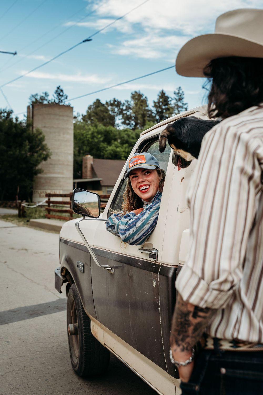 Smiling woman in baseball cap leans out vintage truck window chatting with person in cowboy hat on rural road.