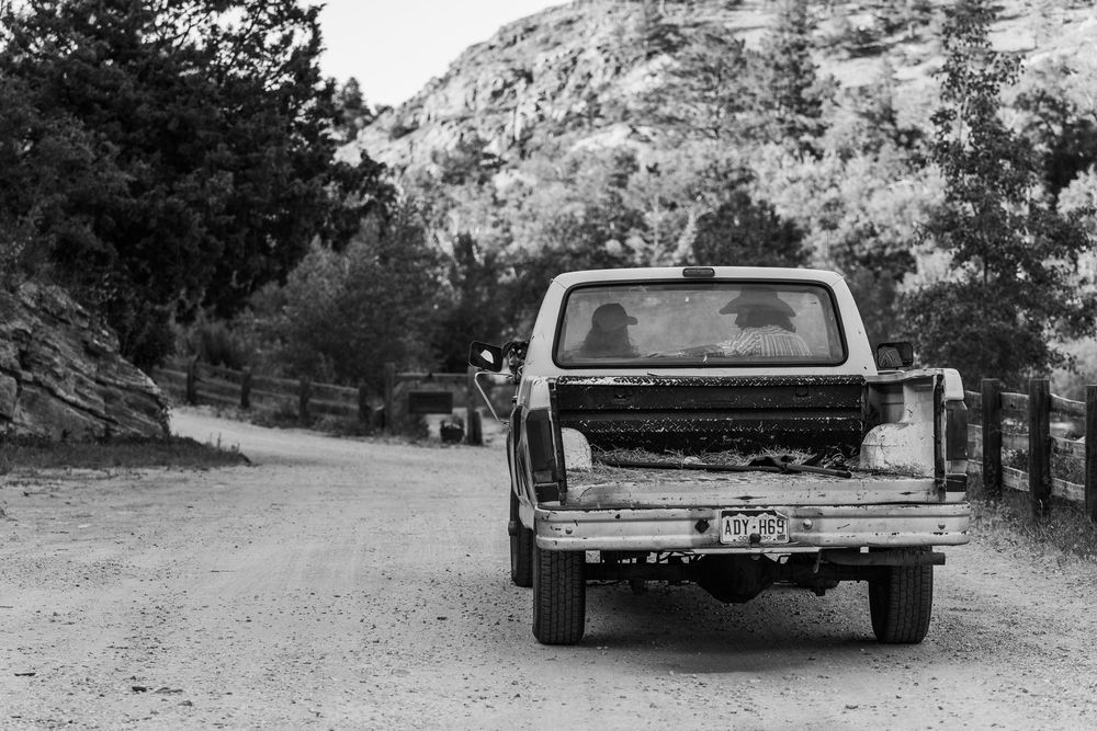 Black and white photo of an old pickup truck parked on a rural dirt road with rocky mountains and trees in the background.