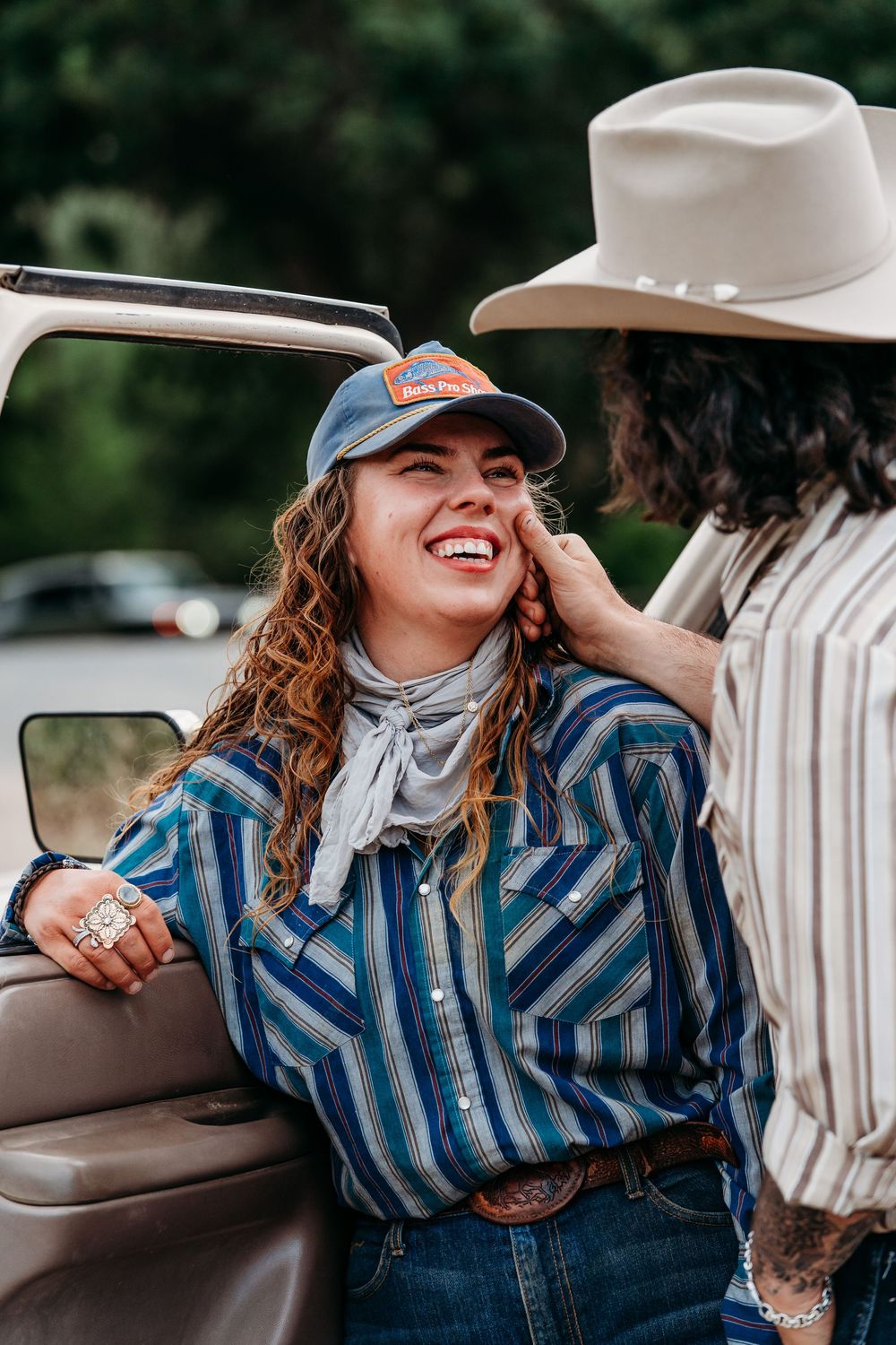 Smiling woman in striped western shirt and trucker hat leans against truck while companion touches her face.
