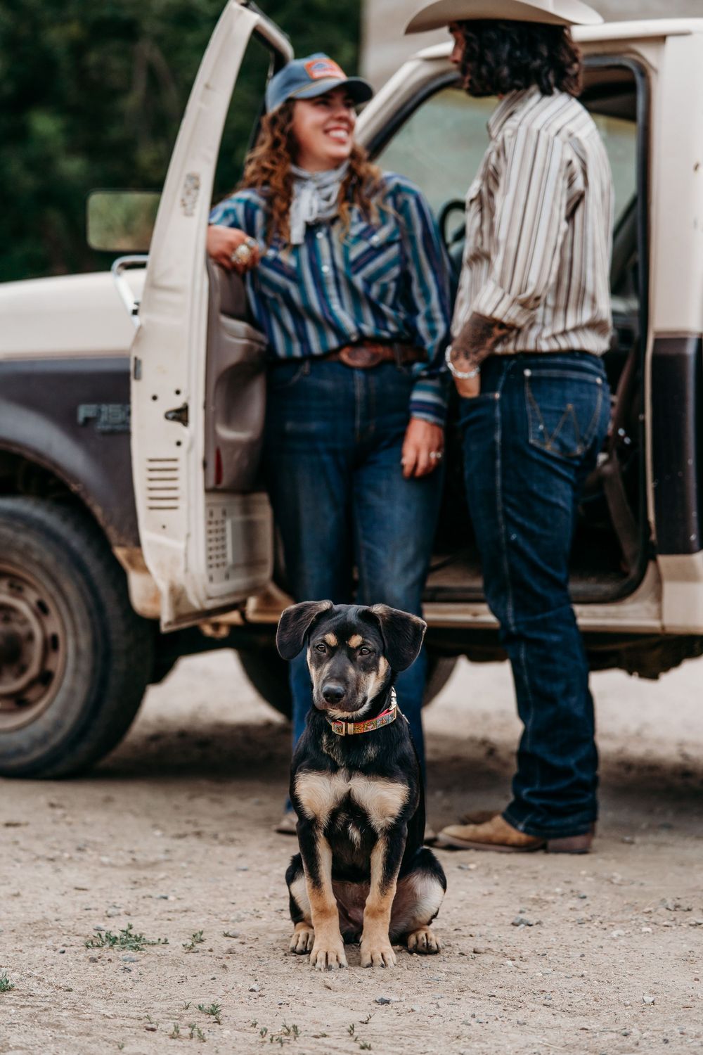 A black and tan dog sits in the dirt in front of two cowboys chatting by a white Ford pickup truck.