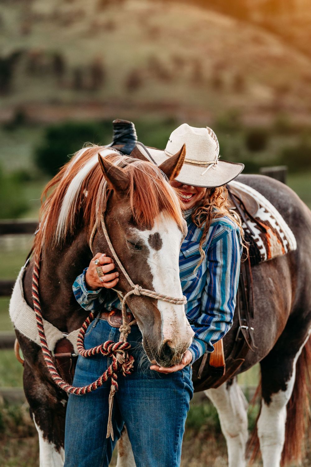 A cowgirl in a white hat and plaid shirt affectionately embraces a brown and white paint horse outdoors.