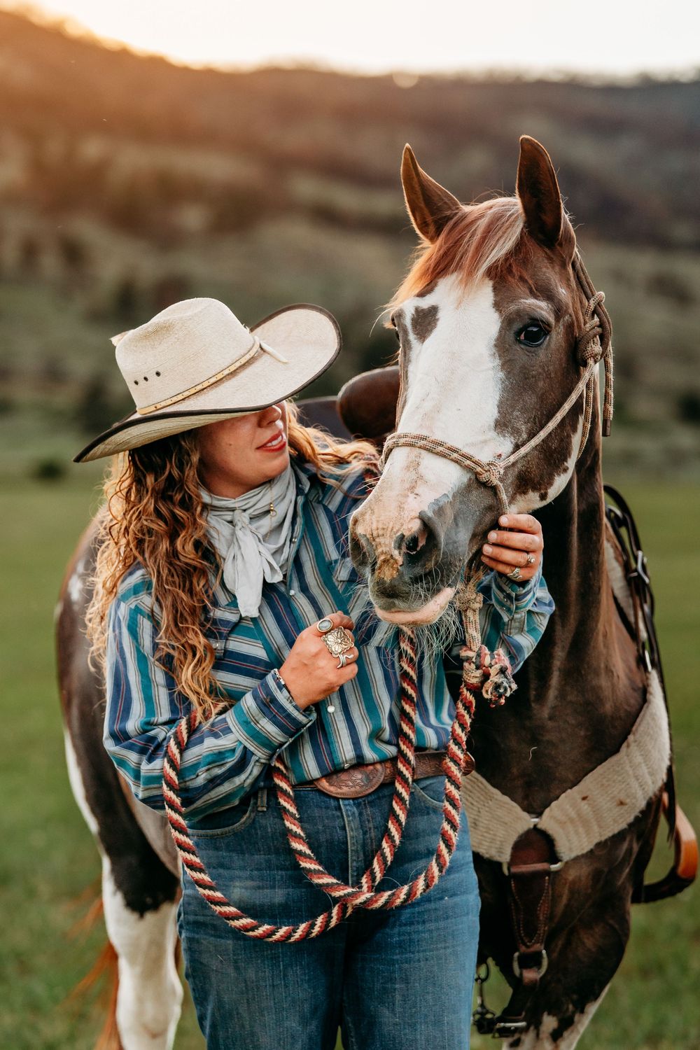 A cowgirl in a striped shirt and white hat tenderly holds the reins of a paint horse at golden hour.