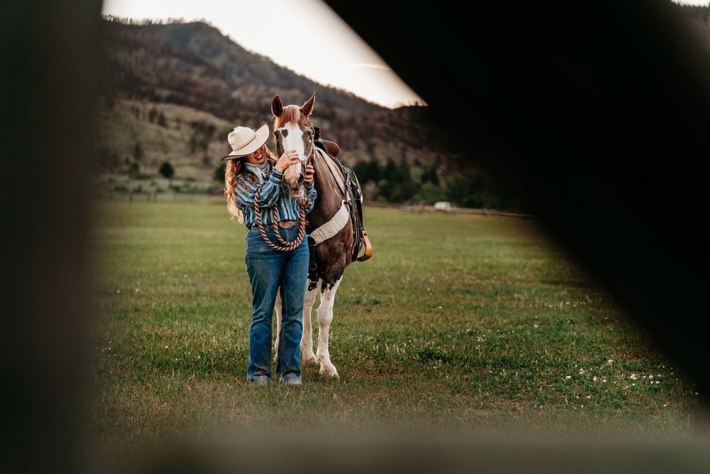 A woman in a cowboy hat stands with a brown and white horse in a lush green mountain pasture.