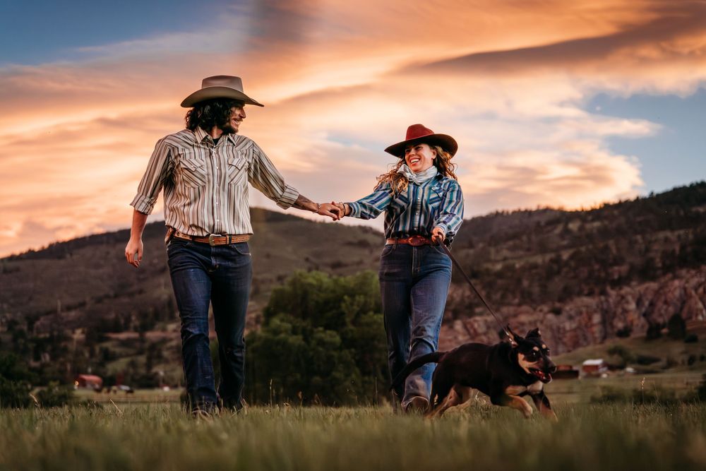 Two cowgirls in western shirts and hats walk hand-in-hand through a meadow at sunset with a black dog.