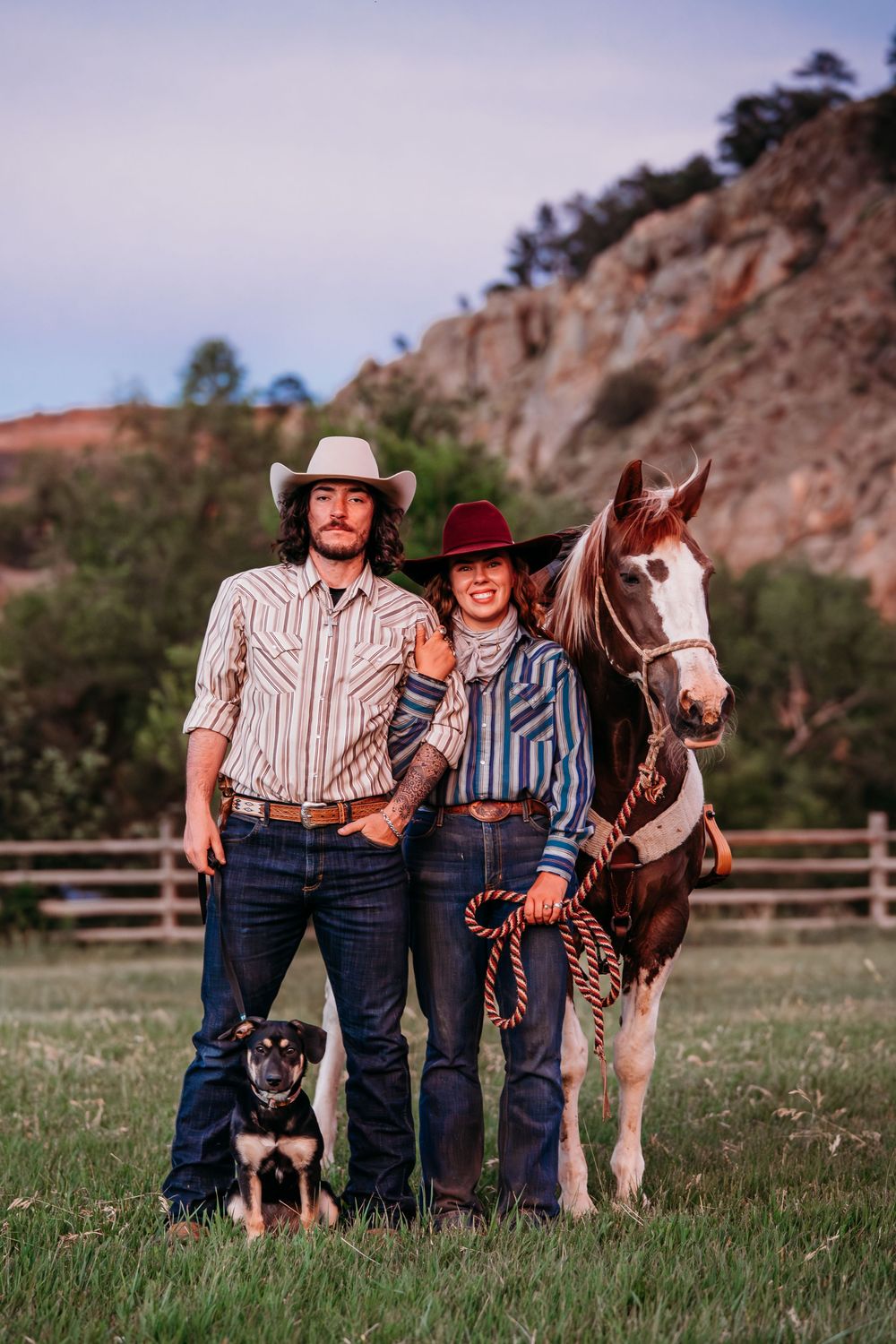 A cowboy couple in western attire pose with their paint horse and dog in a scenic ranch setting.