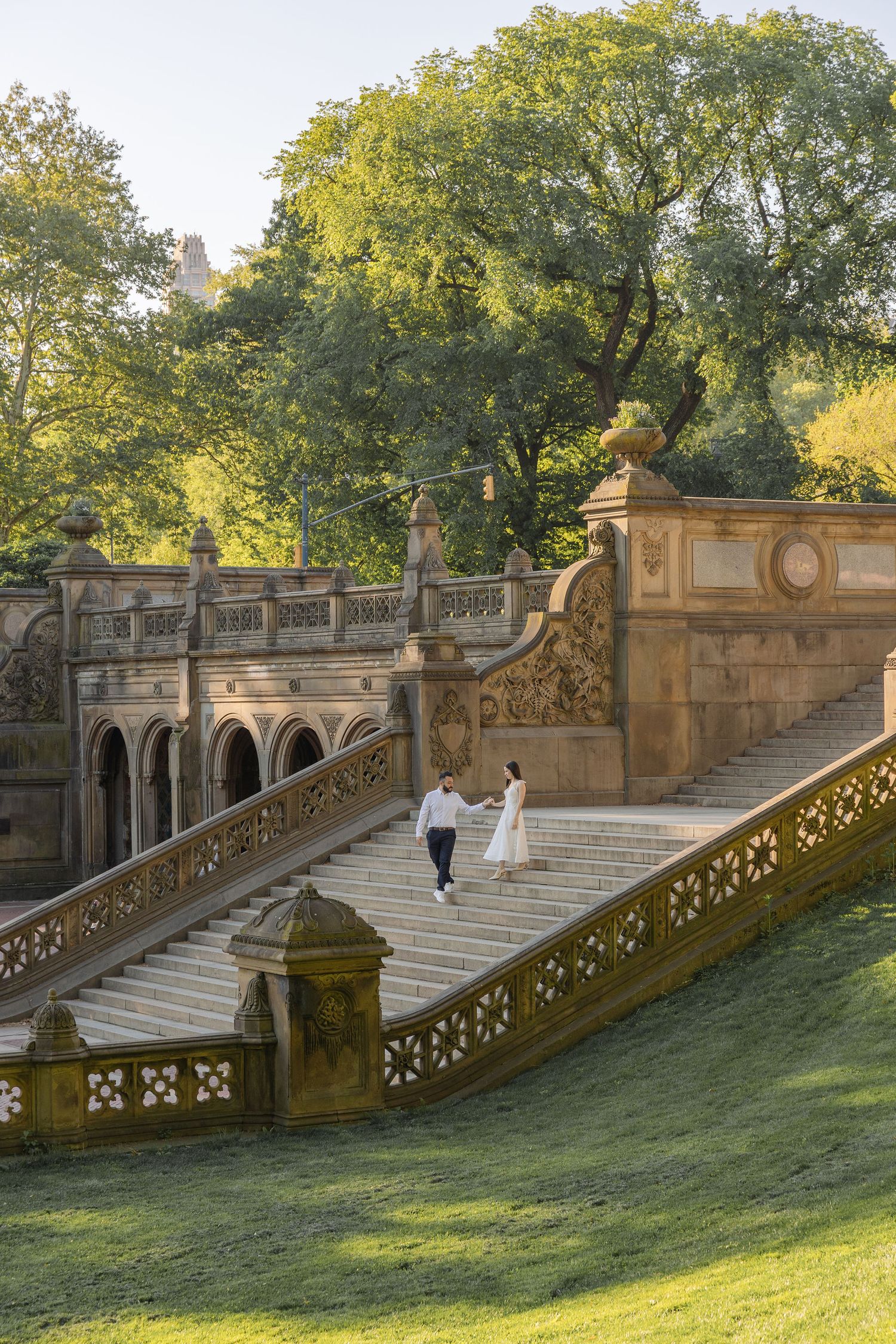 Couple in wedding attire walks hand-in-hand on Bethesda Terrace steps in Central Park, surrounded by lush green trees.