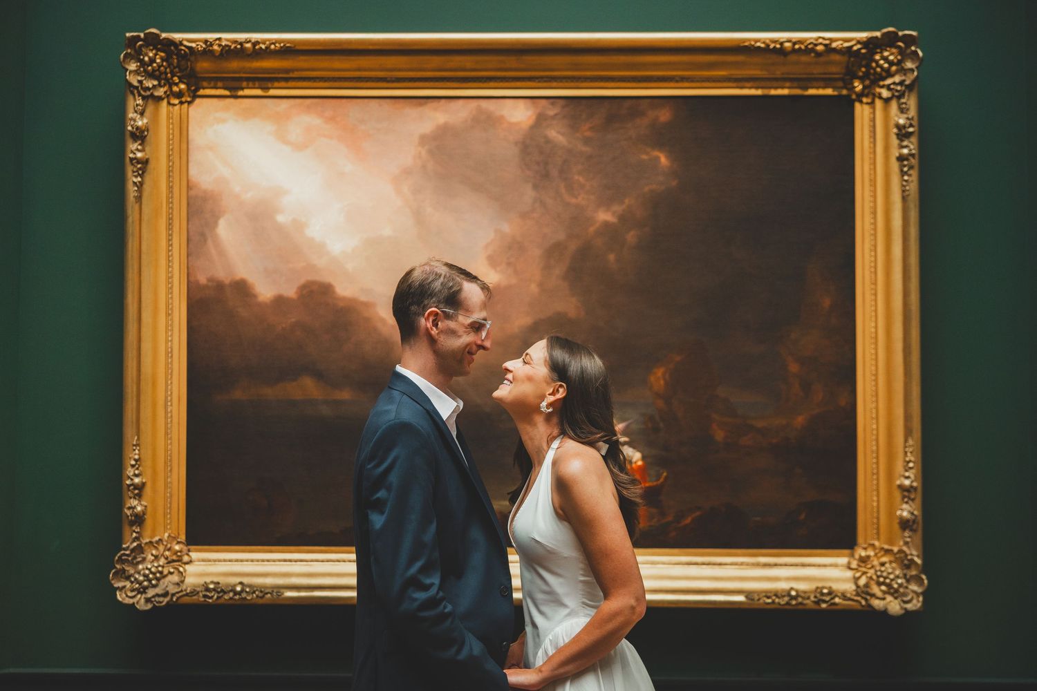 Couple sharing a romantic moment in front of a large ornate gold-framed painting in an art museum.