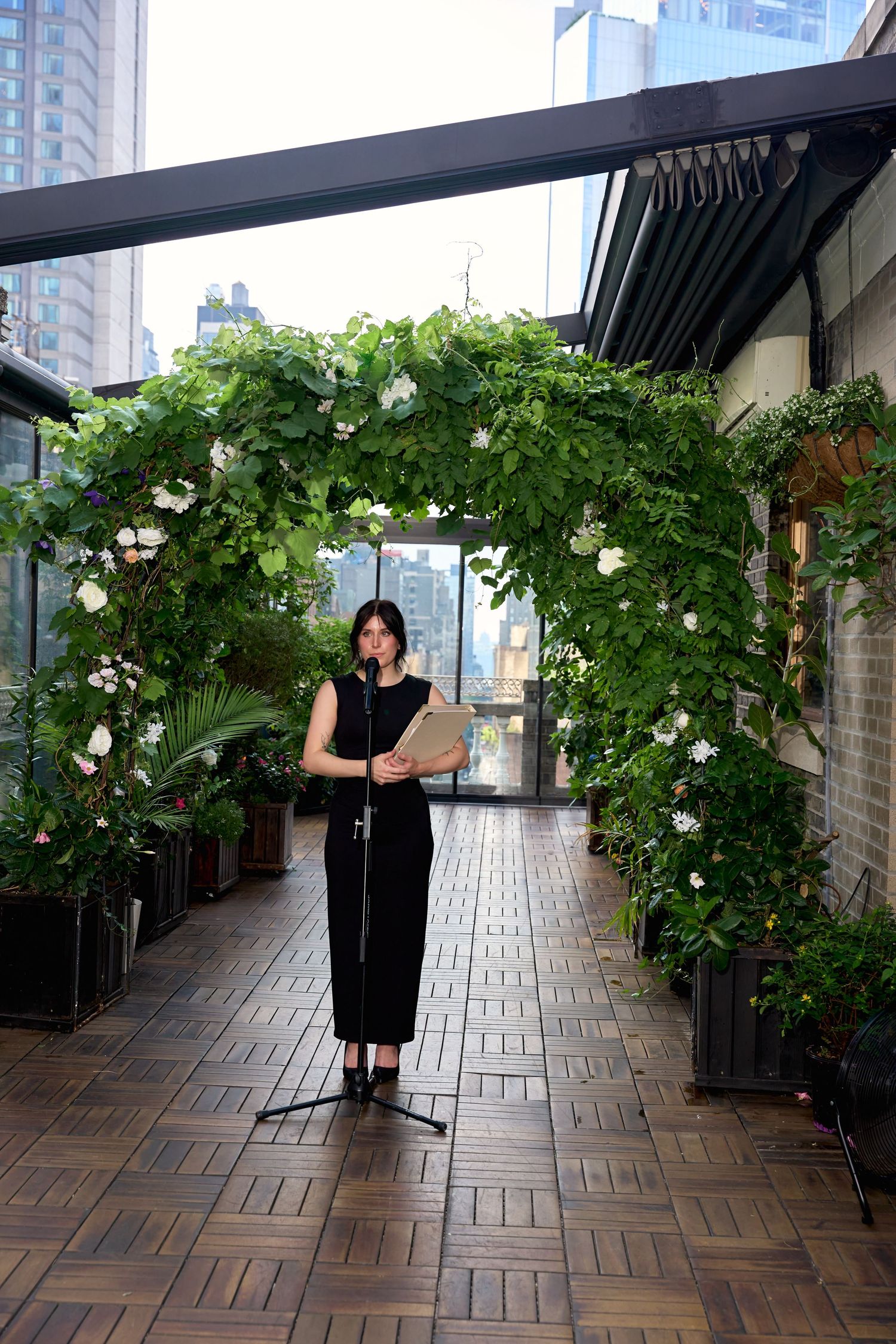 Woman in black dress reads at microphone under floral arch on urban rooftop terrace with city skyline backdrop.