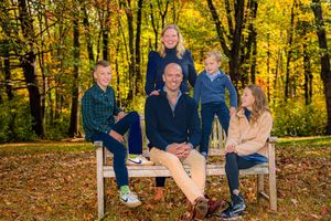 Family of five posing on a wooden bench surrounded by vibrant autumn foliage in a scenic outdoor park setting.