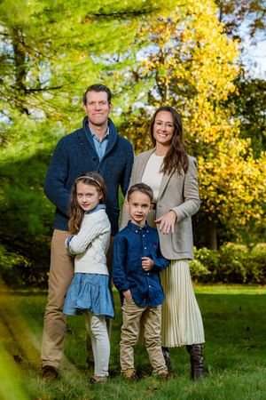 Family of four posing outdoors in autumn, parents and two children smiling among vibrant fall foliage.