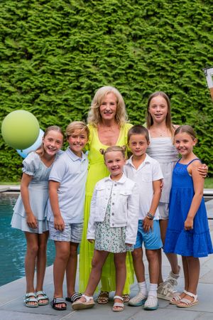 A smiling blonde woman in a green dress poses with six children near a pool with a lush green hedge backdrop.