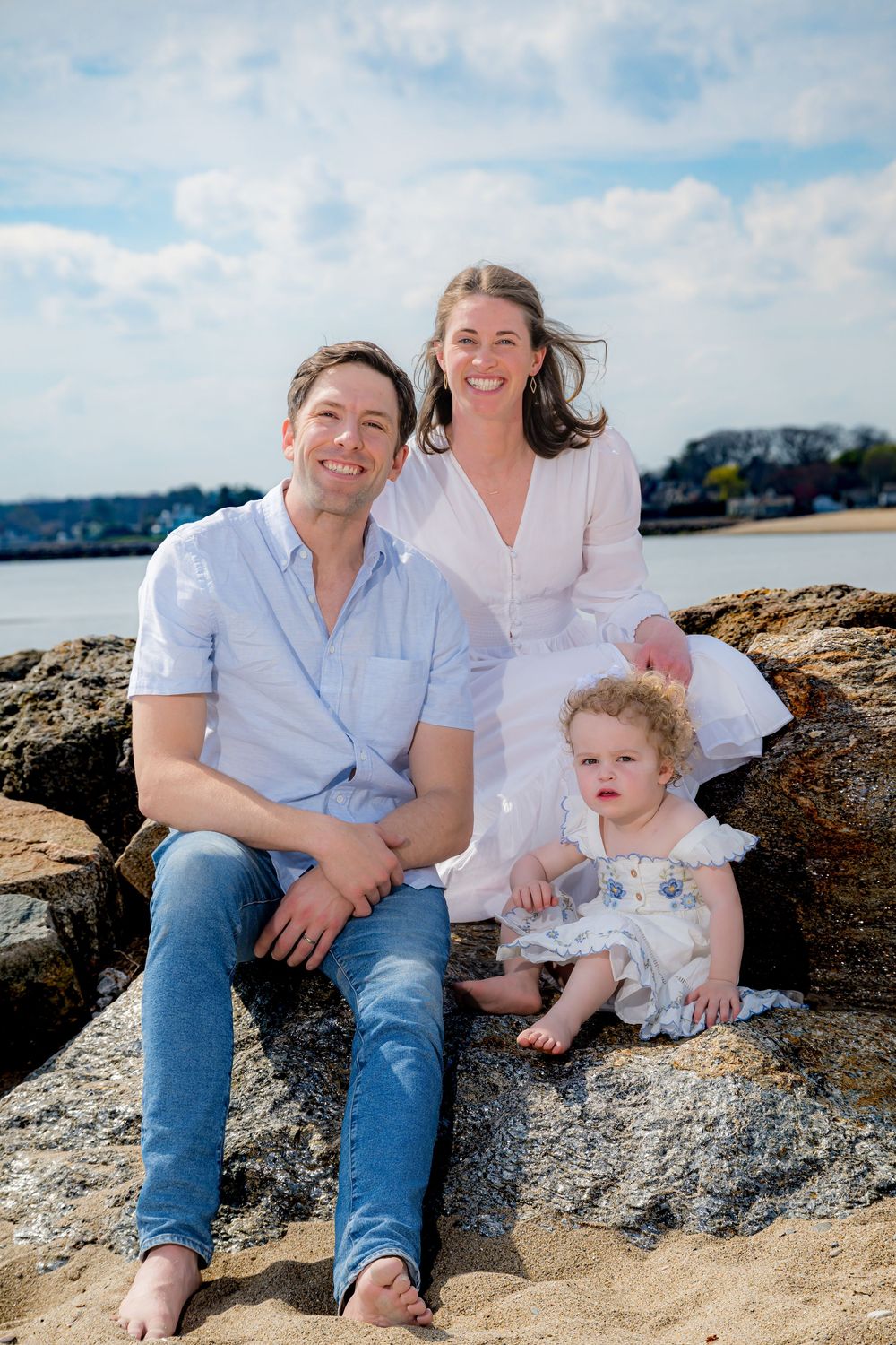 A happy family of three sits on rocky beach, parents smiling with curly-haired toddler, ocean backdrop on sunny day.