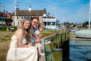 Couple with toddler posing on a marina dock in wedding attire on a sunny day near a waterfront building.