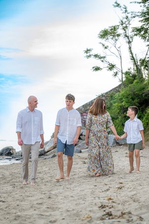 Family of four walking barefoot on a sandy beach, wearing white outfits, with lush green trees in the background.