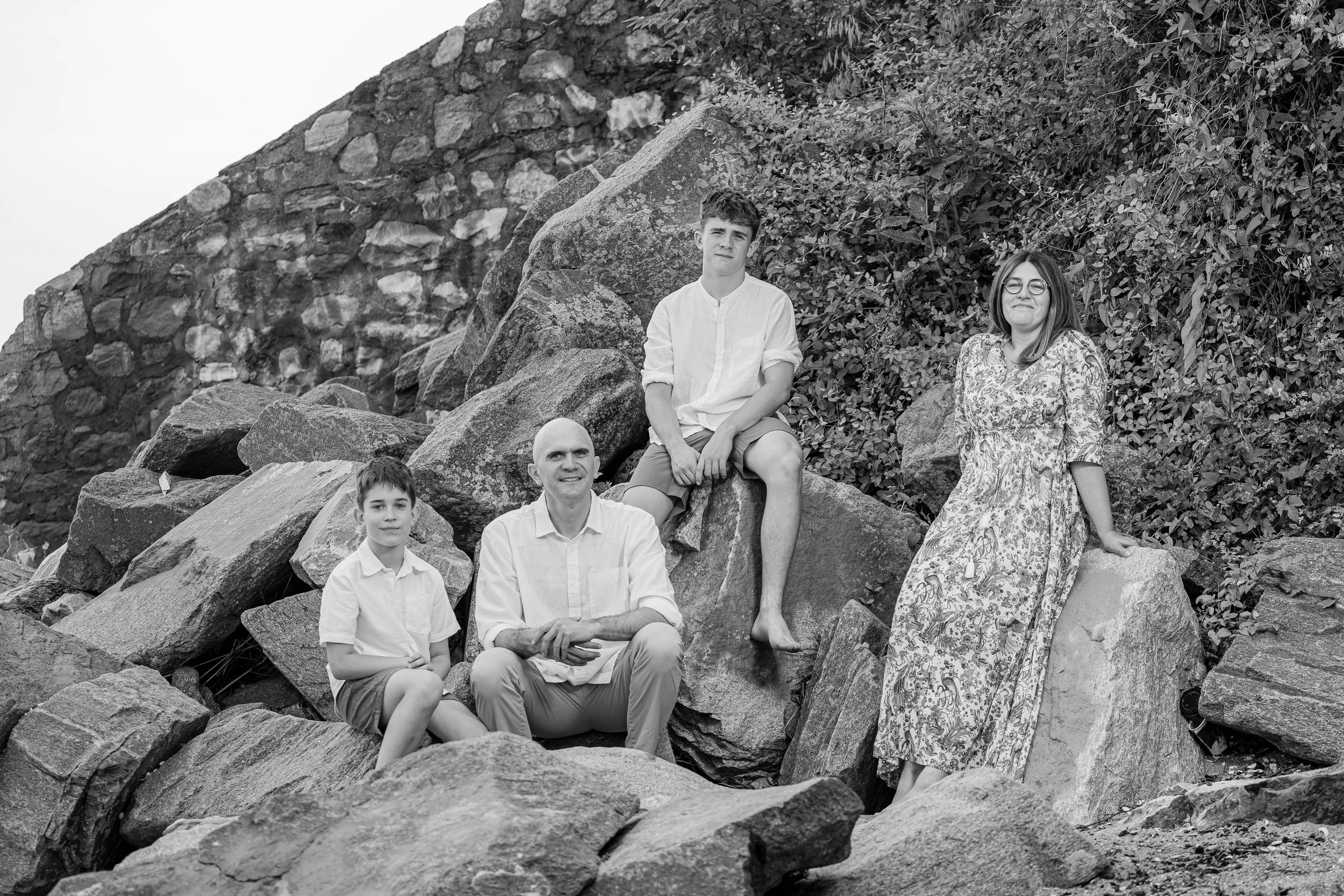 Black and white family photo of four people posing on rocky shoreline with stone wall backdrop.