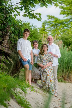 Family of four posing outdoors near a fallen log surrounded by lush greenery on a sandy path.