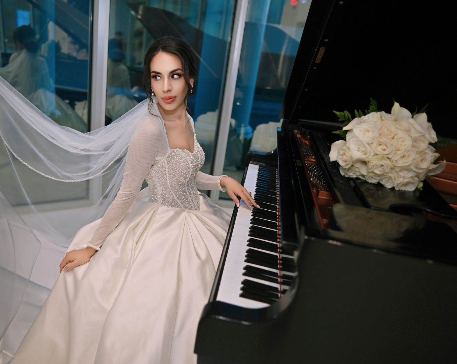 Elegant bride in a white gown and veil seated at a grand piano with white roses bouquet.